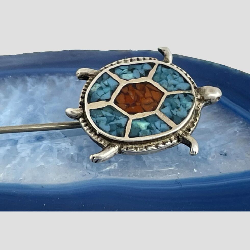 A close up photo of the mosaic turquoise turtle. Its oval back is separated into 6 sections with a central hexagon. Each section has silver dividers giving a very neat look. The central hexagon has red coral mosaic which the 6 sections around it have mosaic turquoise. With its head at the tip, tail opposite end and 4 flipper feet spread out , it appears as though swimming especially against the blue of the agate.
