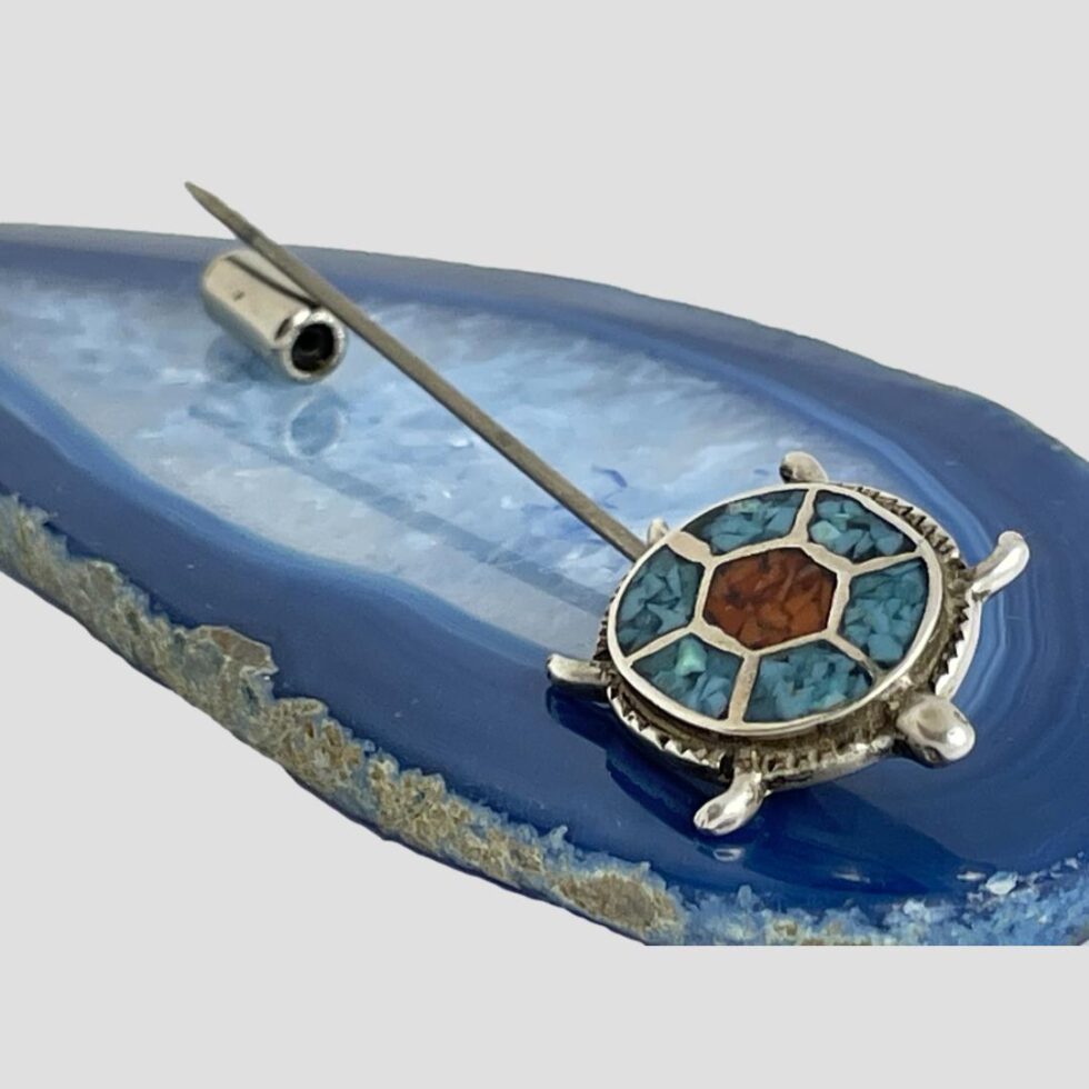 Photo of the mosaic turquoise turtle pin still on top of a slice of agate this time with the turtle in the foreground and bottom right corner. The pin has its safety cover removed and laid next to the exposed pin in the top left corner background.