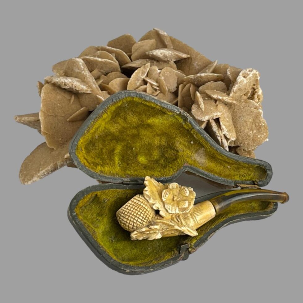 Main photo of antique Victorian pipe displayed inside its case with the mouthpiece just sticking out over the edge. The open lid of the case is being propped open by a rock of desert sand rose. The inside is moulded and covered in olive green velvet. Although the end of the pipe is slightly sticking out over the end, it is clearly visible that a segment of the pipe is missing. This would be the clay bowl where the tobacco goes. It should be possible to find a replacement or make a new bowl.