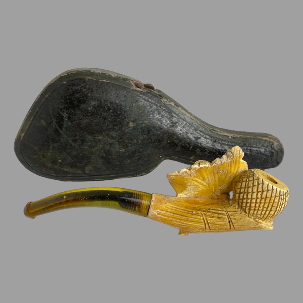 Photo of Victorian pipe displayed with the lesser decorative side on show. The pipe is laid horizontally in front of its case which is propped up head down.