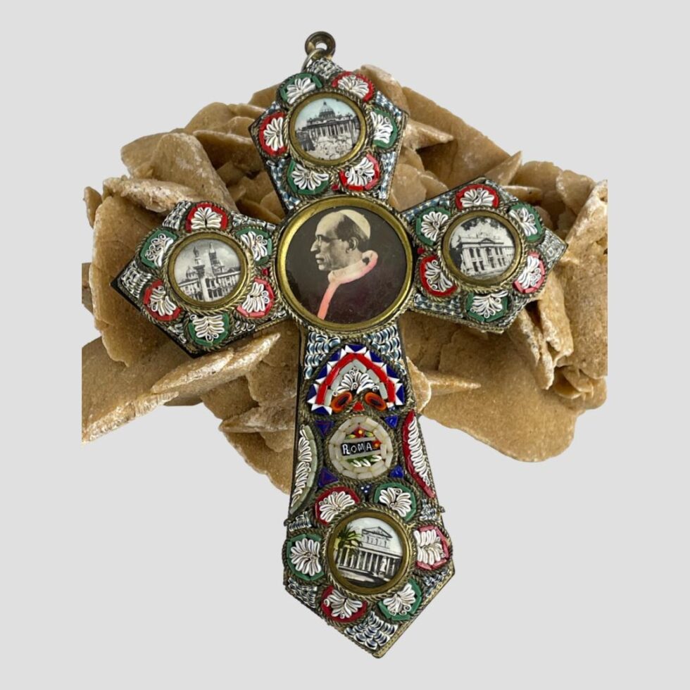 Main photo of micro mosaic papal cross displayed leaning against a rock of natural desert sand rose with the cross front on display. The mosaic colours are in red, white, blue and green with round brass open locket areas at the tips with pictures of famous Roman landmarks and in the centre is the largest picture with Pope Pius XII. The collar of the popes cloak has been hand coloured a pale pink which appears a little faded with time. The mosaic is in good order and nearly all present.