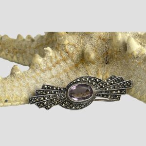 Main photo of the silver marcasite amethyst brooch displayed in front of a starfish and forward facing. The overall shape of the brooch is like a stylised bow, the oval cut amethyst laid horizontally at centre with 5 graduating marcasite studded rows. There is also a sweep over of marcasite to the top and bottom of the amethyst. Simple and very stylish.