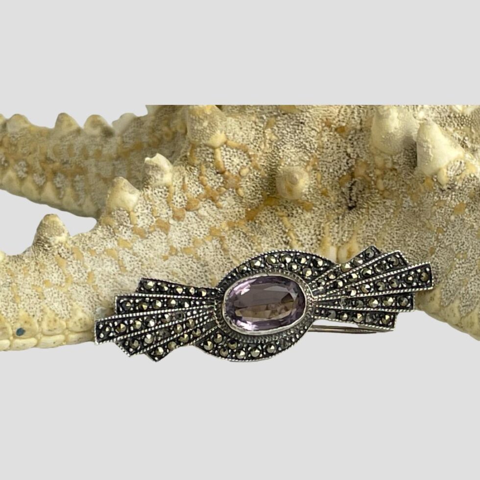 Main photo of the silver marcasite amethyst brooch displayed in front of a starfish and forward facing. The overall shape of the brooch is like a stylised bow, the oval cut amethyst laid horizontally at centre with 5 graduating marcasite studded rows. There is also a sweep over of marcasite to the top and bottom of the amethyst. Simple and very stylish.