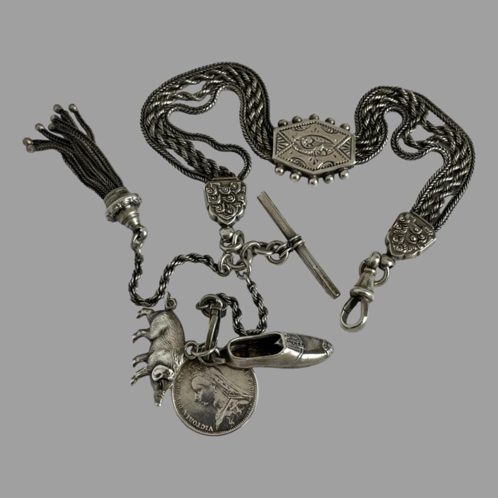 Photo of the Victorian silver Albertina chain displayed on a flat surface against a grey background. The chain has 4 strands (2 outer rope chain and inner 2 twist rope) and a flat slider which has 5 silver balls to each side and has an unusual engraved design akin to abstract floral. The dog clip is on the right and on the left we have the tassel at left, T-bar at right and in the centre the large round clasp with the 3 charms.