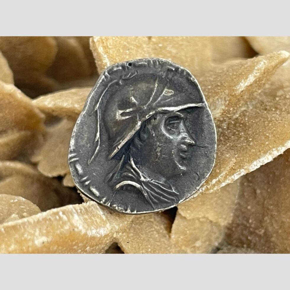 Main photo of Greco-Bactrian one Drachma coin showing the obverse with image of Eucratides I in profile. He is draped and wearing a Boeotian helmet with bulls horn and ear. It is displayed on top of some desert sand rose which can be seen in background. In remarkable condition for an item of this age - just goes to prove, they don't make them like they used to!