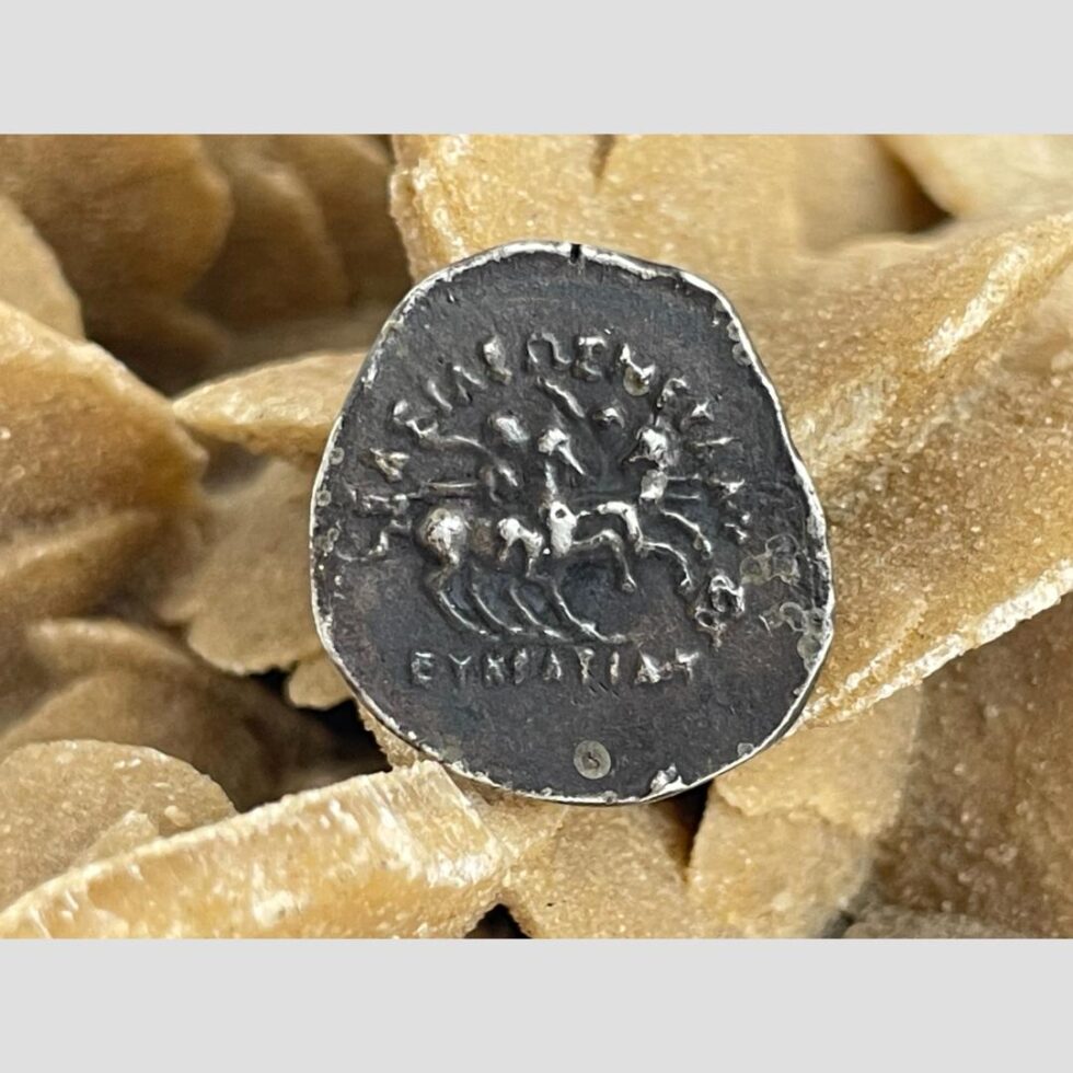 Photo of the reverse of Greco-Bactrian one Drachma coin showing Dioskouri on horses. The image is a little worn but can still make out the men on horses. The desert sand rose is still in the background.
