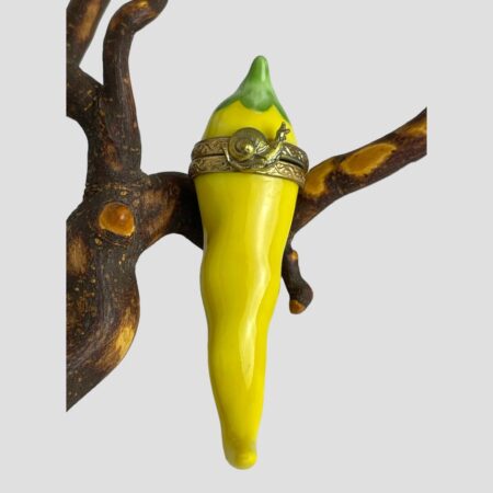 Main photo of Limoges yellow chilli box displayed leaning upright on a twig with the opening side with snail on display. Beautifully hand-painted and looking very real if it wasn't for the brass band at the top with the snail. Green topped and tapered to a chilli point at the end, this is a mild chilli of a lemon yellow colour.