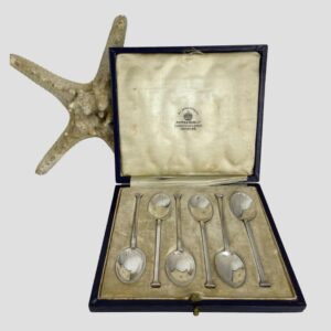 Main photo of Art Deco silver teaspoons displayed inside their original fitted case. The open lid shows the Mappin & Webb stamp to the silk which has some small rips and foxing. The spoons are laid top to tail in fitted velvet covered slots. There is part of a starfish peeking out from behind the open lid to the left for display purpose only.