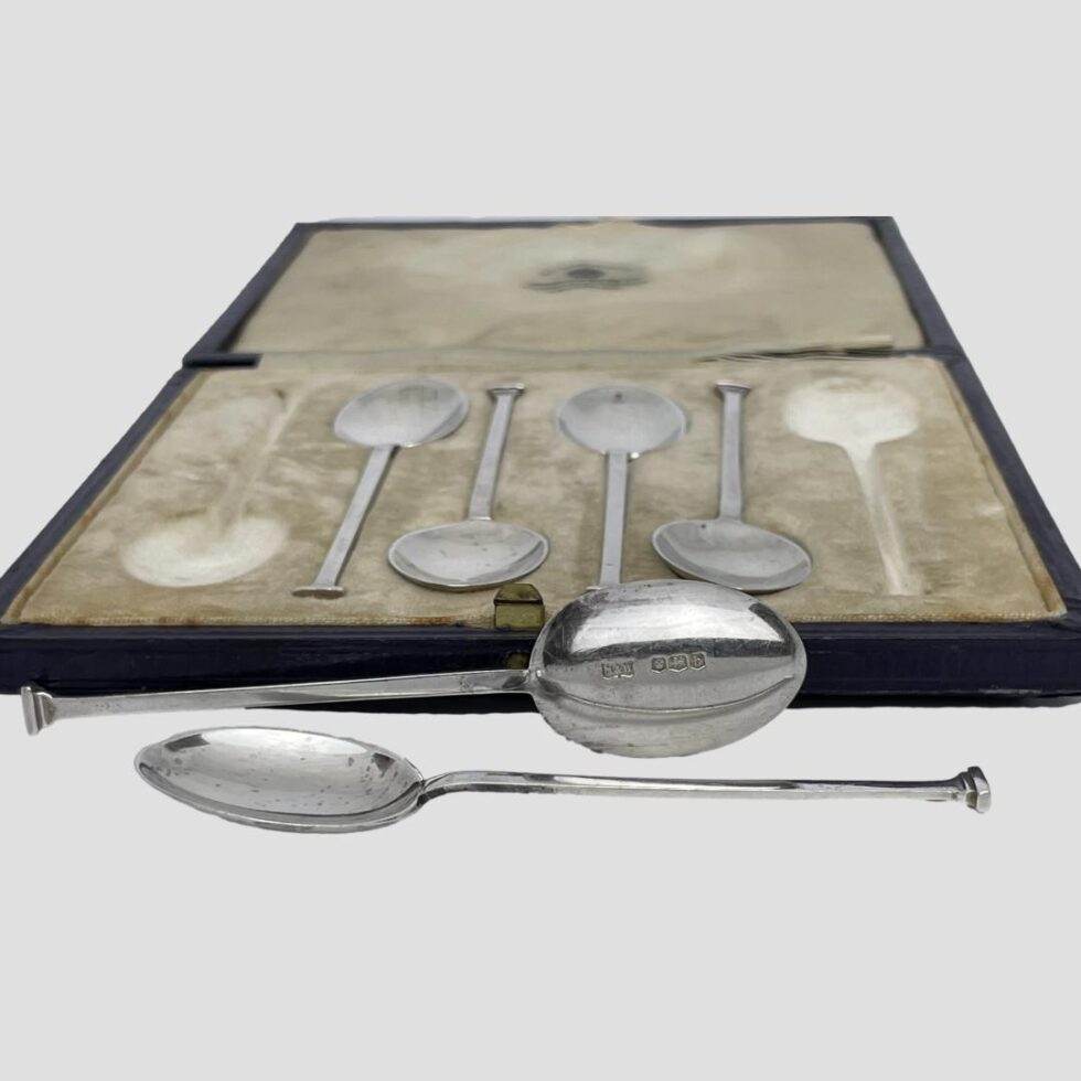 Photo, from an eye level, 2 spoons have been removed and laid in the foreground of the case. One is laid flat with the bowl on the left. The other is horizontal but upright with the back of bowl on display and to the right with the full hallmark. The case is wide open with the lid flat down.
