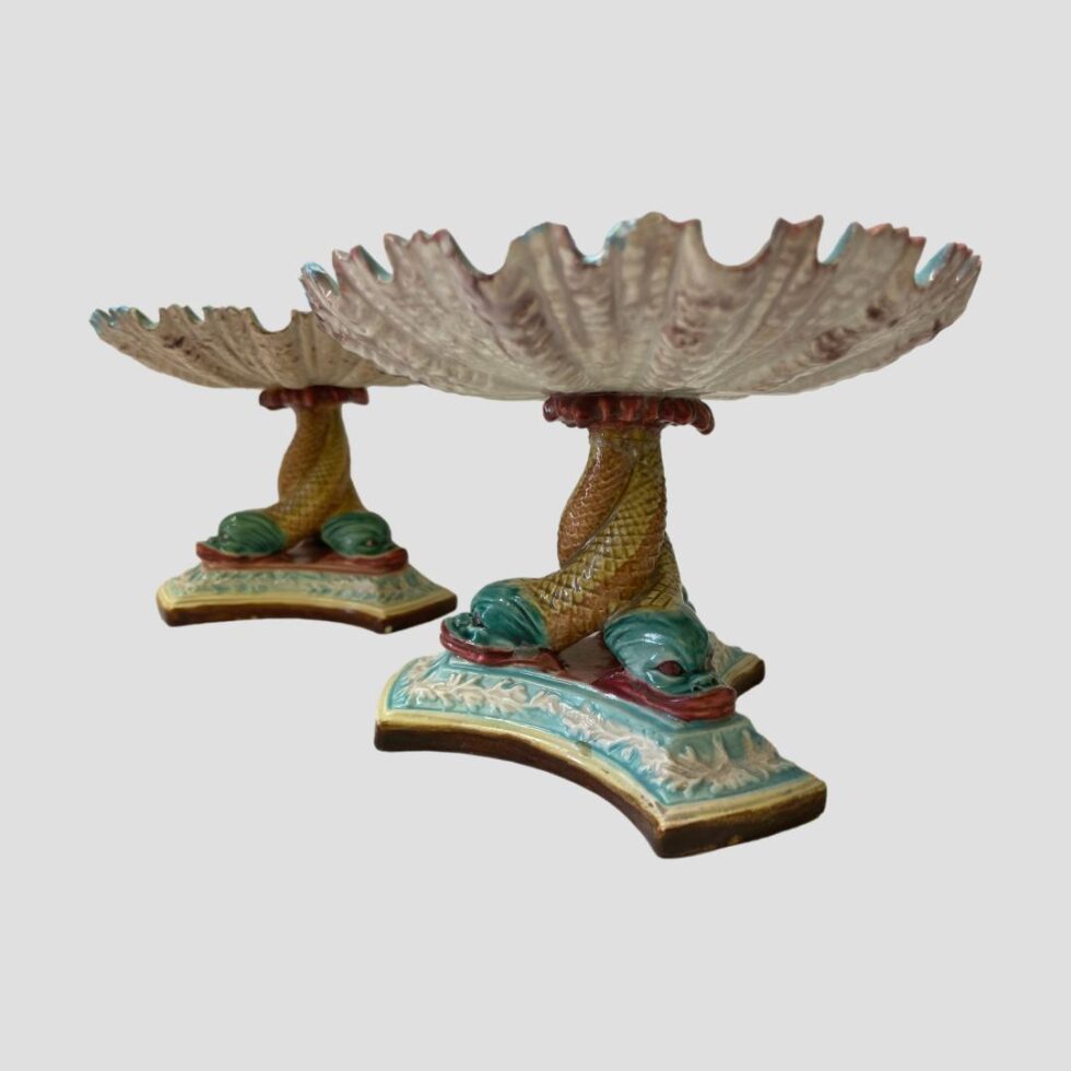 Photo of Art Nouveau sweetmeat dishes seen from eye level with one dish in front of the other and to the right. The triple dolphin column and shell bowl detail is clearly visible.