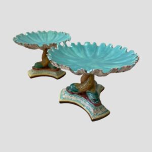 Main photo of the 2 Art Nouveau sweetmeat dishes with one in front of the other and off slightly to the right. We are looking diagonally downward with the turquoise blue inside of the bowls in clear view. The triple footed columnwith dolphins is visible to the dish on right only (other is obscured by one in foreground)
