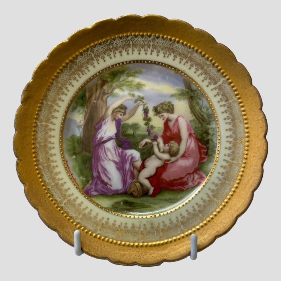Close up photo of neo classical style plate with the 2 ladies playing with a baby. The lady on left in purple dress is holding aloft a vine with grapes . The lady in red on the right has the baby in her lap who is holding onto the bunch of grapes on the end of the vine. There are a few small specks of wear with the main one at the base of the woman in reds dress.