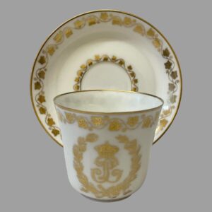 Main photo of antique Sêvres Cup &Saucer with cup placed in front of the saucer which is displayed upright on a stand. The decorated side of cup (opposite end to handle) is shown. The 2 sides do not have any decoration other than the frieze of ivy leaves around the lip.