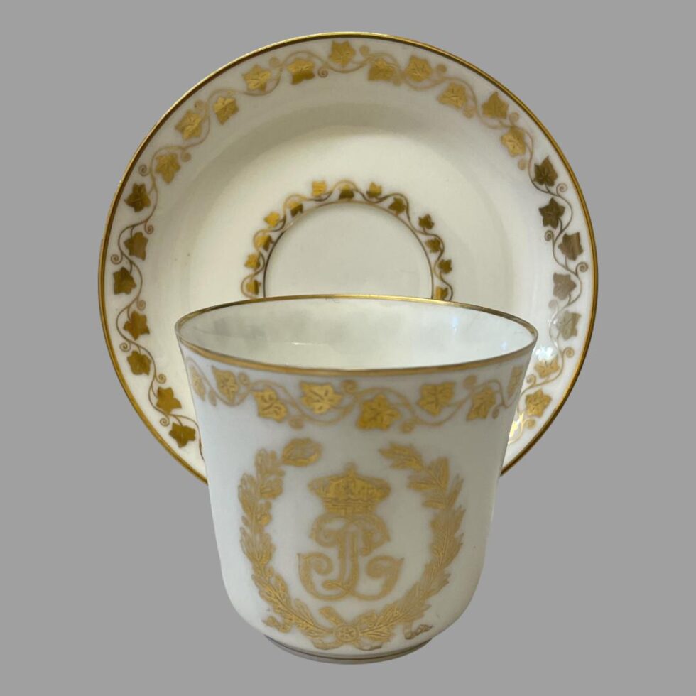 Main photo of antique Sêvres Cup &Saucer with cup placed in front of the saucer which is displayed upright on a stand. The decorated side of cup (opposite end to handle) is shown. The 2 sides do not have any decoration other than the frieze of ivy leaves around the lip.