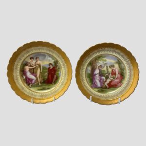Main photo of Neo classical style plates displayed upright and side by side on a white plate stand. On the left a group of 4 women either preparing to or having just bathed in the nearby river and on the other, 2 ladies playing with a baby. Both plates have a gilt outer rim followed by a cream coloured section with classical gilt design. The images are in a variety of colours with natural skin tones and rural scenes and colours of the clothing are bright like raunchy red or passionate pink. Both scenes from a period long ago yet somehow very nostalgia inducing.