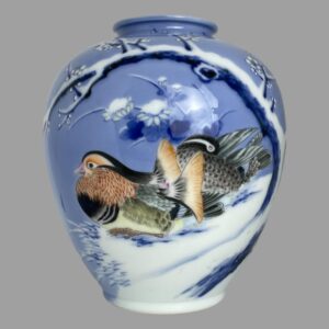 Main photo of Fukagawa Seiji vase displayed with the mandarin ducks side on show. The colourful male with its fine plumage in orange, yellow and pink along grey and black is to the left whilst the less colourful female sits behind pruning her grey and black feathers. The pair are sat on snow covered ground beneath a curved branch of white winter plum in blossom - a symbol of endurance and impending arrival of spring. The base colour of the vase is a beautiful pale blue with darker patches with the winter plum tree in a darker blue with snow covering.