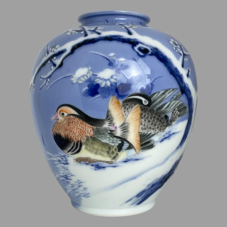 Main photo of Fukagawa Seiji vase displayed with the mandarin ducks side on show. The colourful male with its fine plumage in orange, yellow and pink along grey and black is to the left whilst the less colourful female sits behind pruning her grey and black feathers. The pair are sat on snow covered ground beneath a curved branch of white winter plum in blossom - a symbol of endurance and impending arrival of spring. The base colour of the vase is a beautiful pale blue with darker patches with the winter plum tree in a darker blue with snow covering.