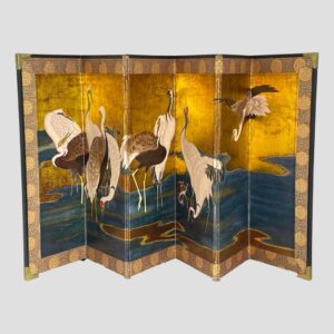 Main photo of Kano Tanyũ Cranes Byobu displayed spread out and forward facing showing the famous scene with cranes wading in a lake. There are a total of 11 cranes, 1 being a colt (baby crane). 10 of the 11 birds are in the water with one on the far right flying into land. The background is mostly golden with the lake a light ink blue with flow lines giving the water movement. The cranes are mixed in colour from the all white to white headed with brown body and of course the little colt which is mostly brown still. All except the colt have a red spot on the heads. The outer edge of the screen has a silk band and the corners are reinforced with brass.