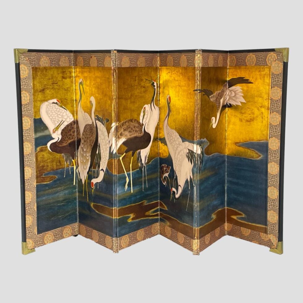 Main photo of Kano Tanyũ Cranes Byobu displayed spread out and forward facing showing the famous scene with cranes wading in a lake. There are a total of 11 cranes, 1 being a colt (baby crane). 10 of the 11 birds are in the water with one on the far right flying into land. The background is mostly golden with the lake a light ink blue with flow lines giving the water movement. The cranes are mixed in colour from the all white to white headed with brown body and of course the little colt which is mostly brown still. All except the colt have a red spot on the heads. The outer edge of the screen has a silk band and the corners are reinforced with brass.