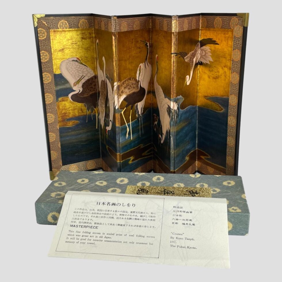 Photo of cranes byõbu displayed open and behind the original box which is laid horizontally in front of the screen. The original paper detailing the piece is leaning against the box. It reads, in the English translation on the left "This fine folding screen is scaled print of real folding screen which was great art in old Japan. It would be good for interior ornamentation not only ornament but memory of your travel' and on the right ' "Cranes" by Kano Tanyü. 17C. The Fukui, Kyoto.'