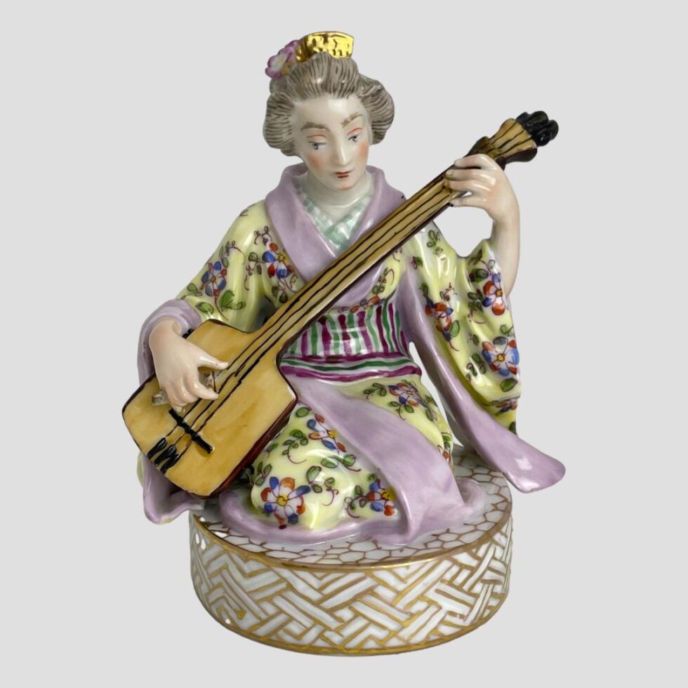 Main photo of Dresden geisha with shamisen displayed with the geisha forward facing. She is sitting in seiza position (on her knees with buttocks on heels) Her kimono is very colourful and not traditional Japanese pale green and white striped under kimono covered with a yellow kimono with red white and blue flowers and green foliage and pale pink outer areas (collar, sleeves etc) Her obi(belt sash) has vertical green cerise and white stripes at the top and a short area beneath with horizontal lines in same colours. She is a right handed player so the shamisen neck is held in her left hand with the body to the right and her right hand holding the bachi (large plectrum)