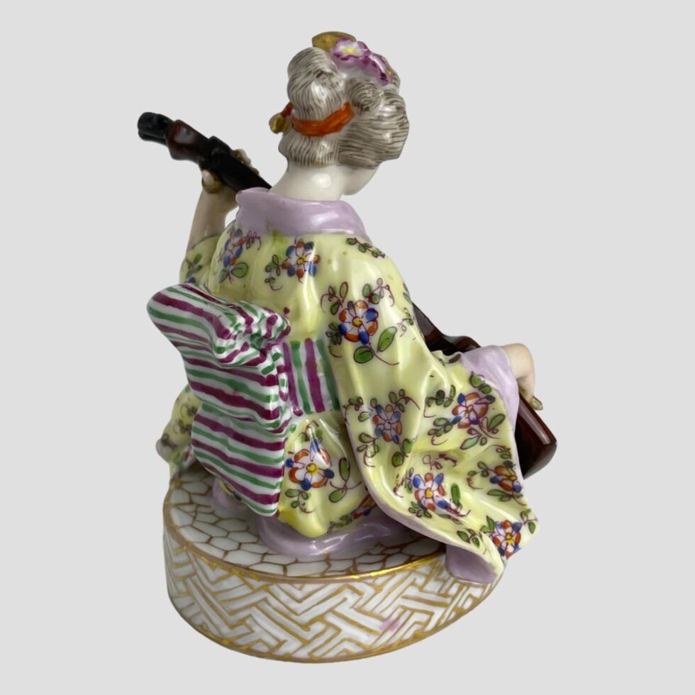 Photo of geisha playing shamisen this time facing away from camera and towards the top right corner. The back of the obi is fully visible and looks like an old humbug sweet with its colours and shape. There is also a red band holding up the bun in her hair at the back. Also just visible is the thumb loop at the back of the shamisen tenjin (headstock on a guitar) She has her thumb through the loop to hold the shamisen sao (neck) steady.