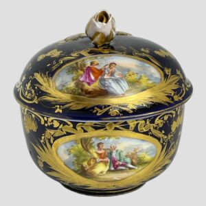 Main photo of Meissen small lidded pot displayed from a near eye level with courting couples scene shown to both lid and pot base. Both scenes show a young couple in brightly coloured Georgian fashion wear (bouffant dresses for the ladies and waistcoats, jackets & breeches for the gentlemen) seated in a rural or garden setting. The base colour of the pot in a dark royal blue with extensive gilt work throughout including around the hand-painted images, enhance the dreamy romantic scenes. The chips to the rose finial and the gilt wear, also around the rim of the lid is visible.