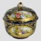 Main photo of Meissen small lidded pot displayed from a near eye level with courting couples scene shown to both lid and pot base. Both scenes show a young couple in brightly coloured Georgian fashion wear (bouffant dresses for the ladies and waistcoats, jackets & breeches for the gentlemen) seated in a rural or garden setting. The base colour of the pot in a dark royal blue with extensive gilt work throughout including around the hand-painted images, enhance the dreamy romantic scenes. The chips to the rose finial and the gilt wear, also around the rim of the lid is visible.