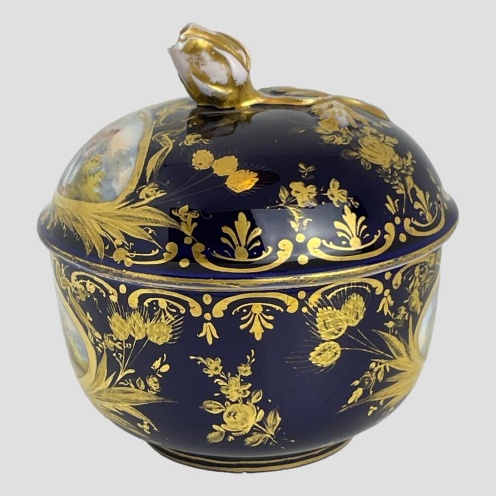 Photo of small Meissen pot showing one side with the 'windows' of romance paintings only just visible to the extreme left and right on the pot bottom and left on the lid. This is a view of the dark royal blue base colour and the incredibly detailed hand-painted gilt work with floral details and swags. The gilt wear on the rose finial is very visible here.