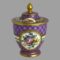 Main photo of 19th century lidded pot seen from an eye level and displayed with the window of different floral bouquet on show and the small window of pink rose and yellow flowers on the lid. The bouquet of flowers include a large pink rose to the centre with blue/purple daisy like small flowers spray above, yellow and orange flowers to the right and a small short string of fuchsia like flowers along the bottom left. The 'window' is framed in gilt with an outer line of gilt jewels (or dots). Approximately at the mid section of this window, a white band extends round from both sides and connects to the other 'window' opposite. This band also has gilt frame to top and bottom and a design of foliage with small red berries. The lid reflects the same layout but in 2D (flat around the outer rim of lid) The centre of the lid rises like a cake with a golden finial rising to a point at the centre. The main area of the pot and lid is in a dark lilac with criss- cross diamond shaped lines in purple. At the centre of the diamond shape is a small white dot known as oeil-de-perdrix in French (partridge eye). It is a very beautiful piece of porcelain and a delight to behold.