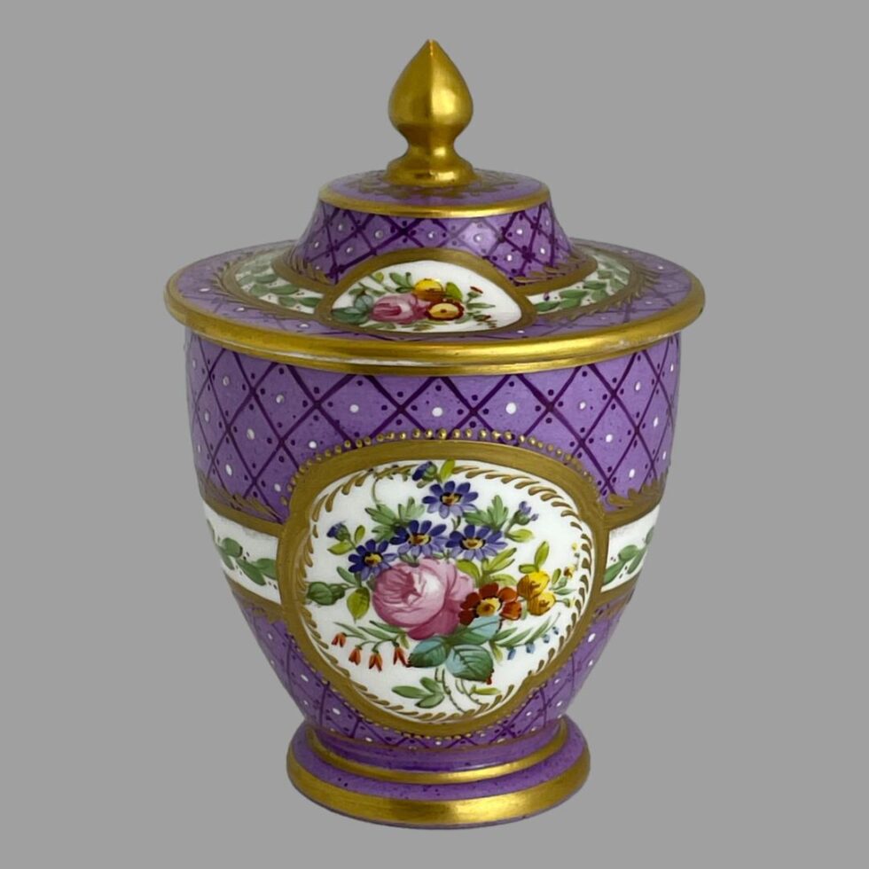 Main photo of 19th century lidded pot seen from an eye level and displayed with the window of different floral bouquet on show and the small window of pink rose and yellow flowers on the lid. The bouquet of flowers include a large pink rose to the centre with blue/purple daisy like small flowers spray above, yellow and orange flowers to the right and a small short string of fuchsia like flowers along the bottom left. The 'window' is framed in gilt with an outer line of gilt jewels (or dots). Approximately at the mid section of this window, a white band extends round from both sides and connects to the other 'window' opposite. This band also has gilt frame to top and bottom and a design of foliage with small red berries. The lid reflects the same layout but in 2D (flat around the outer rim of lid) The centre of the lid rises like a cake with a golden finial rising to a point at the centre. The main area of the pot and lid is in a dark lilac with criss- cross diamond shaped lines in purple. At the centre of the diamond shape is a small white dot known as oeil-de-perdrix in French (partridge eye). It is a very beautiful piece of porcelain and a delight to behold.
