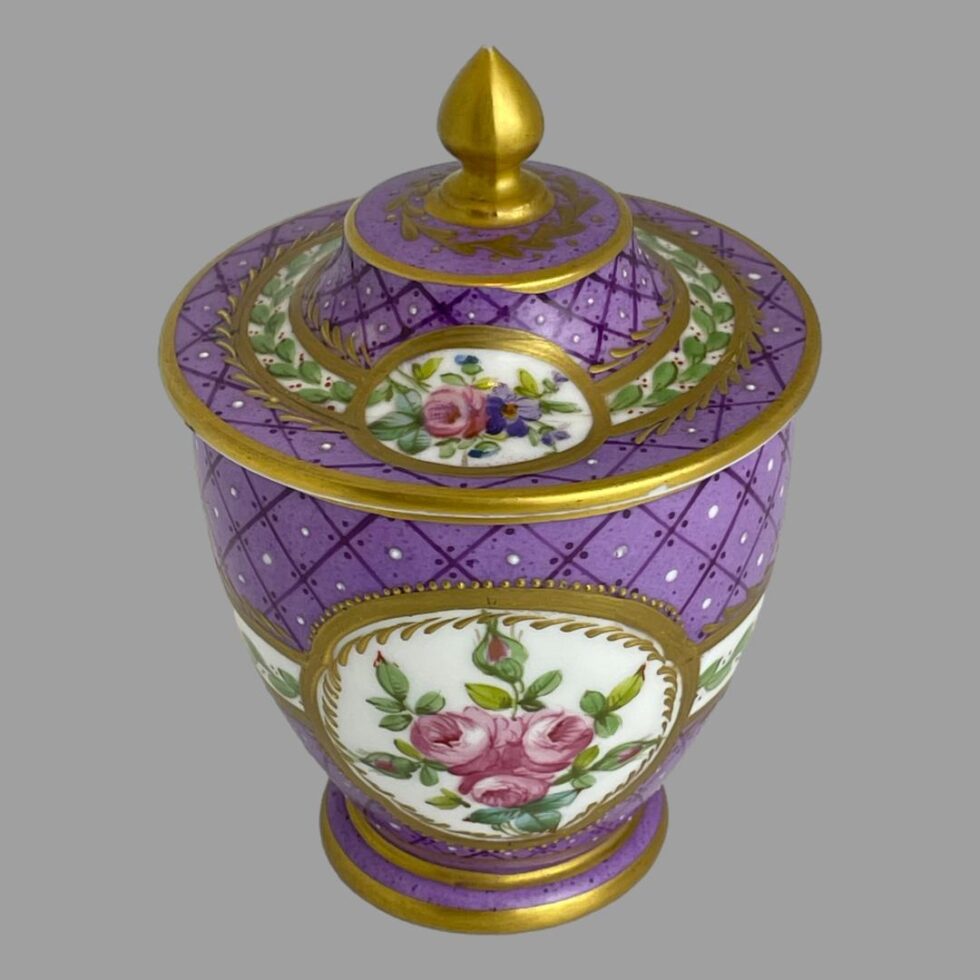 Photo of 19th century lidded pot this time slightly raised and looking diagonally downward showing more of the lid. The opposite side of the windows to main photo are on display. The large window on the pot is a spray of pink roses only while the lid displays a pink rose with small purple flowers.
