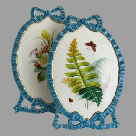 Main photo of pair Minton porcelain pictures displayed with one in front of the other and to the side. The front picture is of the ferns in various shades of green, orange and pink. A red butterfly can be seen top right and a ladybird bottom right of the ferns. The incredible work put into these pictures with the turquoise blue of the ribbon frame ending in a bow at the top and bottom is quite incredible. It is sometimes hard to fathom that these are made entirely of porcelain!