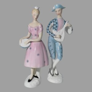 Main photo of Doulton Harlequin &Columbine figurines displayed with both forward facing to camera. Columbine is to the left in a pretty pink dress with purple dandelion seed head decoration. The dress is off the shoulder with short sleeves finishing mid way down her upper arm. A small white crown sits on her head. She holds a tambourine in her right hand with the left hand poised over it as if tapping. The tambourine has a pale blue edge. Harlequin, to the right, is in a pale blue outfit with triangle pattern down the front and down the top of the arms. The underarms of the outfit is in plain white as is his mandolin and waist sash. The jacket or top is open and he has a pale pink vest underneath. He wears a bicorn hat with a white pompom to the right (left in photo) His knee length pale blue pants also have the dandelion head pattern along the side seams. Both are wearing black or grey shoes and standing on a white base.