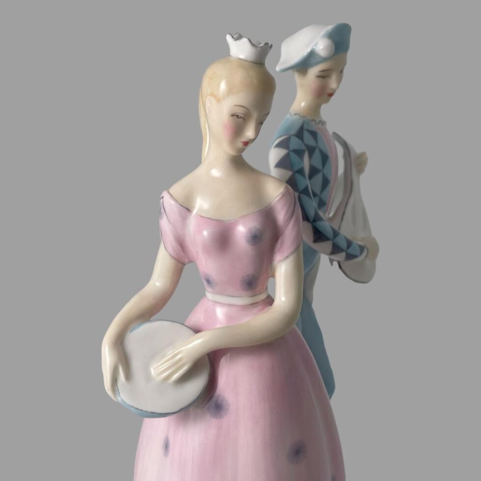 Close up photo of the figurines upper bodies. Columbine is in the foreground , her body is forward facing and her head it tilted downward to the left. Harlequin is in the background with body shown from the side and his head facing downward to the right. With the figurines posed like this it appears as they are both looking at something in the same spot on the ground.