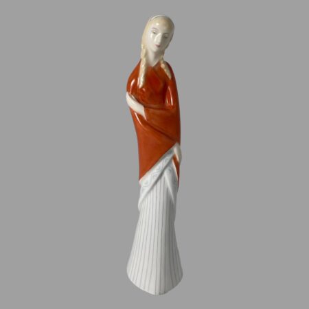 Main photo of Royal Doulton teenager figure displayed forward facing and from an eye level. This is a tall and slender teenager in a dark orange shawl and long white pleated skirt that covers her feet. The shawl is wrapped around and she is holding her right arm above the elbow with her left hand and her right hand crosses over below to her left hip - almost in a cuddle of self. She has pale blonde hair in bunched plaits and a white hair band with blue edges. Very dreamy looking facial expression.