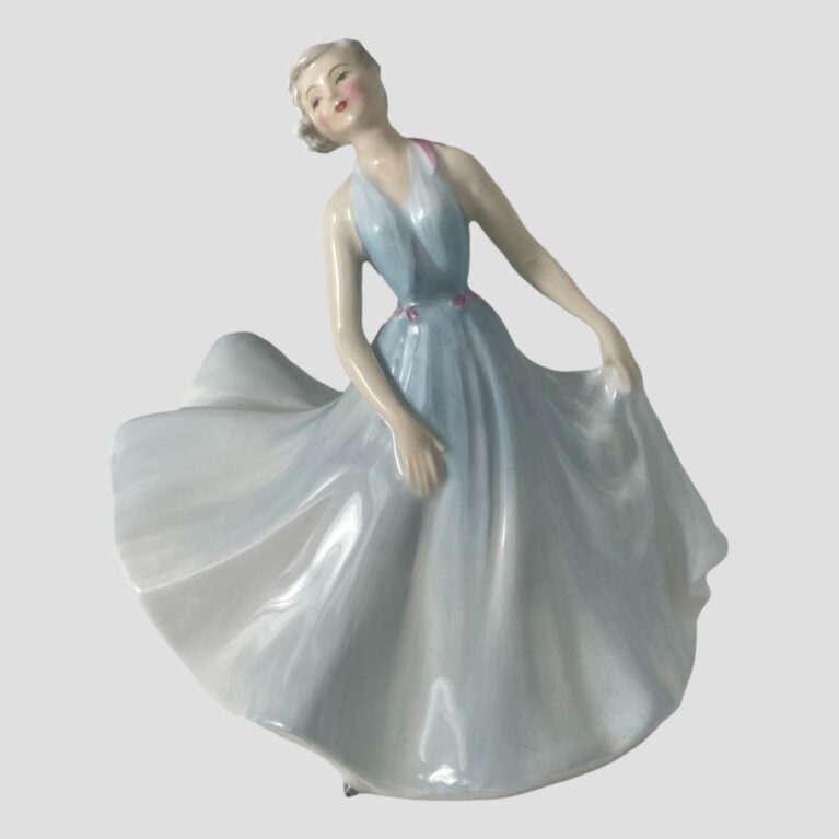 Main photo of Royal Doulton pirouette figure displayed from an eye level and forward facing. The figure has pale porcelain skin, white blonde hair and has rouge and red lipstick on. Her long halter-neck gown is in baby blue shade with pink ribbon accents to the waist and one shoulder. Her dreamy facial features reflect in her pose, holding the skirt of her full dress with the left hand and right arm hanging in front. She is in mid twirl with the full skirt billowing out to the left of photo giving an impression of movement.