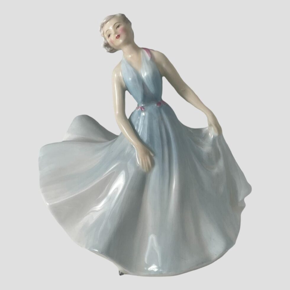 Main photo of Royal Doulton pirouette figure displayed from an eye level and forward facing. The figure has pale porcelain skin, white blonde hair and has rouge and red lipstick on. Her long halter-neck gown is in baby blue shade with pink ribbon accents to the waist and one shoulder. Her dreamy facial features reflect in her pose, holding the skirt of her full dress with the left hand and right arm hanging in front. She is in mid twirl with the full skirt billowing out to the left of photo giving an impression of movement.