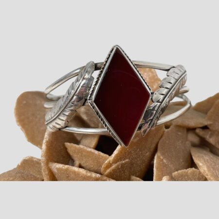 Main photo of red onyx silver cuff displayed on top of a rock of natural desert sand rose with the bracelet front forward facing. The red onyx has a deep rich red colour and is set with an outer frame of sterling silver. The diamond suit shape juts out at the top and bottom points of the diamond. Like the diamond suite in a pack of cards, it is longer vertically. The cuff has 3 silver lines which join at the cuff ends. The 2 silver feathers, one on each side of the red onyx, sit top to tail to each other. One the left, the tip of the feather leans alongside the top lean of the onyx diamond shape and to the right, the feather tip leans alongside the lower lean of diamond shape. The ends of those feathers curl slightly outward and attaches onto the opposite silver band.