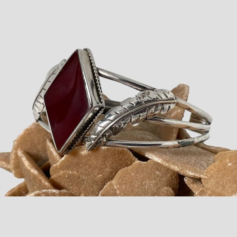 Photo of red onyx silver cuff bracelet still on top of a rock of desert sand rose this time with the onyx facing towards the left. One full silver feather is on show.
