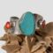 Main photo of Mexican wavy silver cuff displayed on top of a rock of natural desert sand rose and the bracelet front forward facing. The turquoise takes centre stage with its size and vibrant colour. It is an uneven shape, mostly oval with a little section jutting out to the left (in this image) around the middle area. Could be seen as a mittened hand. The stem corals on either side of the turquoise is a vibrant red graduating in shade to a near white centre with a single red dot in the middle. The wavy design gives each stone a platform. A very striking piece of vintage Mexican jewellery.