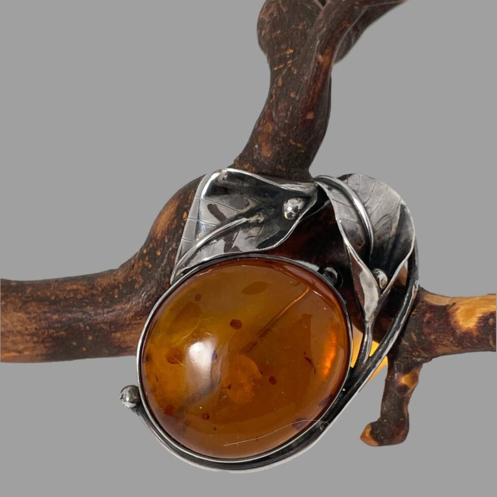 Main photo of Deco style amber brooch displayed leaning against a small twig of cherry tree. The amber cabochon has some inclusions that look like the dots on a ladybirds back and the brooch pin, which is visible through the amber gives an impression of the split of the ladybirds wings. The silver cala lily flower heads are to one side, in this photo to the top right with one above the other so creating a half frame of flowers and the flower stems hug the lower frame of the amber (in this photo)
