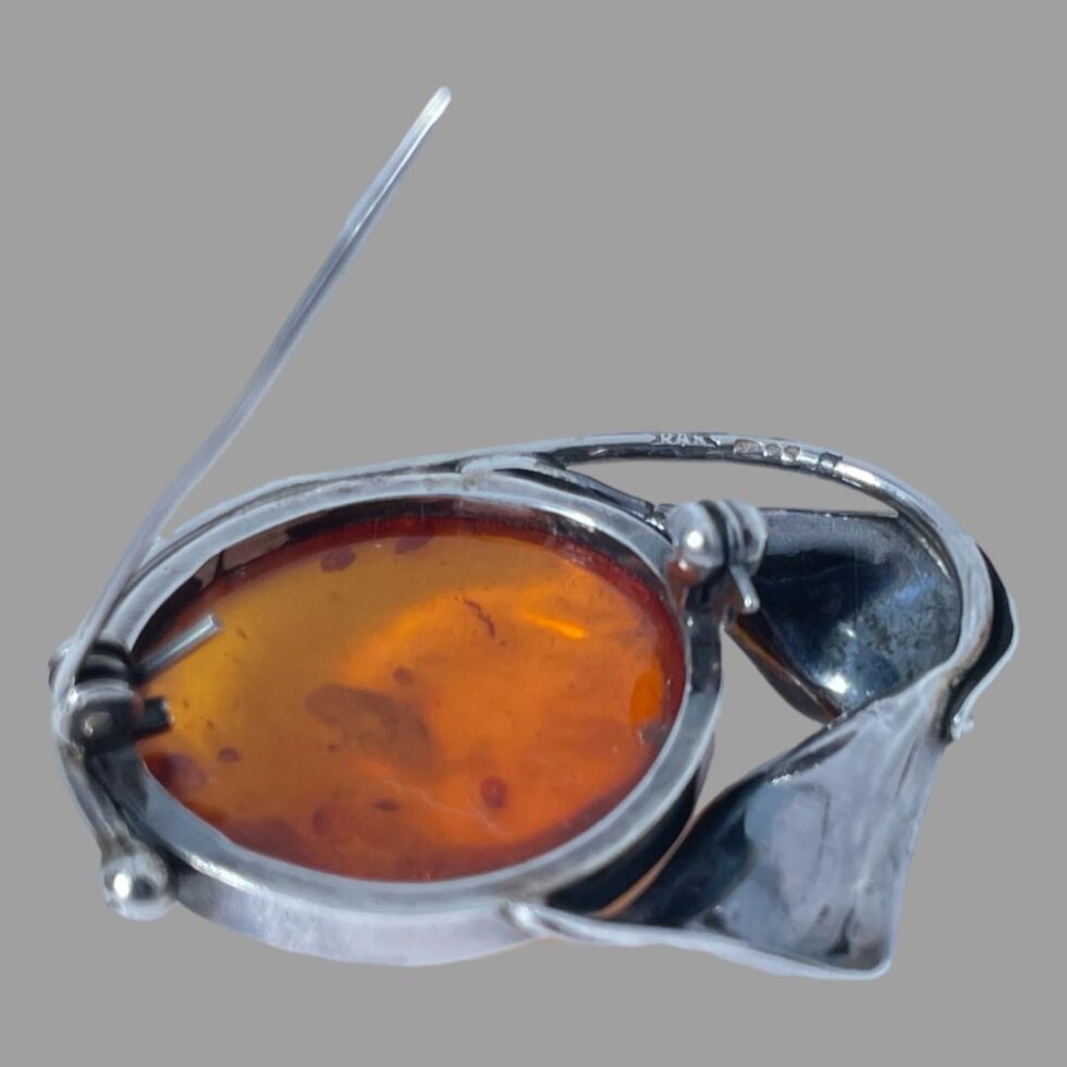 Photo of the back of amber brooch seen with the pin open and leaning upright. The sterling silver of the brooch looks slightly blue tinted here owing to the studio light. The hallmark is visible on the stem of one flower in the top right.