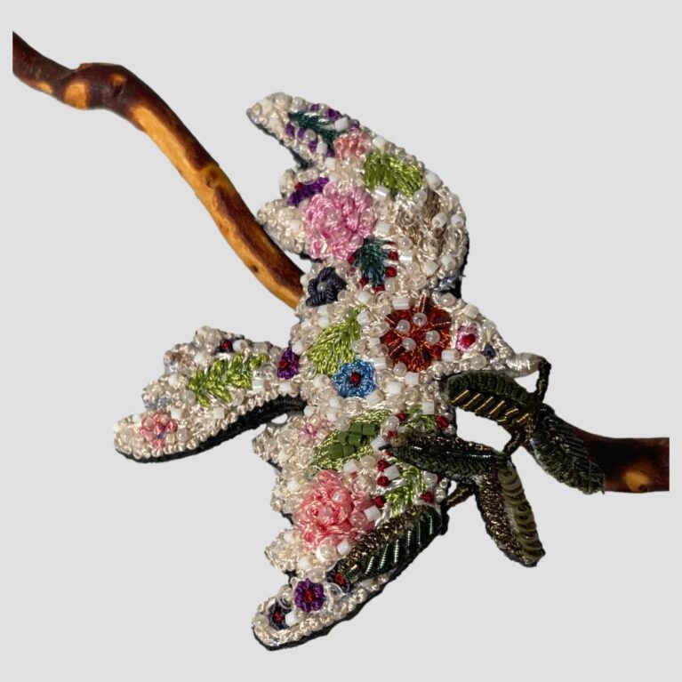 Main photo of trovelore peace dove brooch displayed leaning against a twig of cherry in the background with the dove flying to the right. The beadwork and the embroidery of this piece is exquisite. The dove is a creamy white base colour with embroidered flowers and leaves embellished with different colour bead. It is a thing to bring much joy on beholding. Lovely gift for someone special or yourself!