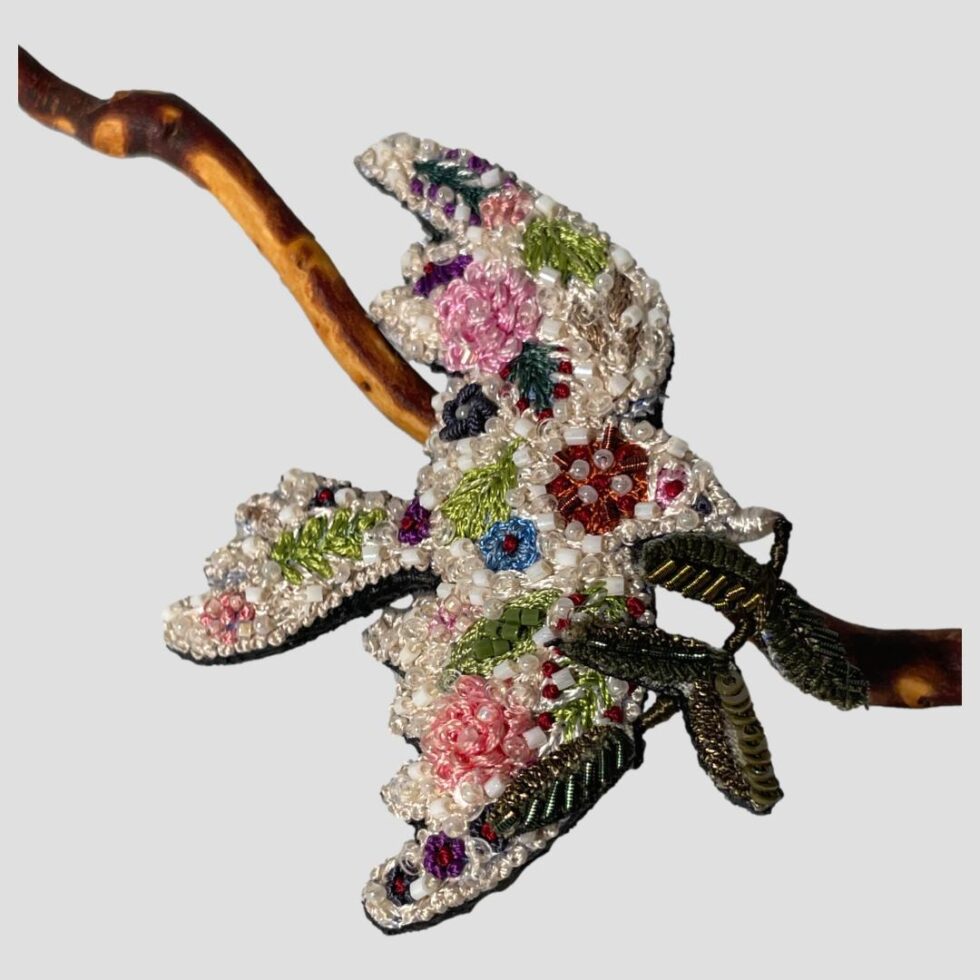 Main photo of trovelore peace dove brooch displayed leaning against a twig of cherry in the background with the dove flying to the right. The beadwork and the embroidery of this piece is exquisite. The dove is a creamy white base colour with embroidered flowers and leaves embellished with different colour bead. It is a thing to bring much joy on beholding. Lovely gift for someone special or yourself!