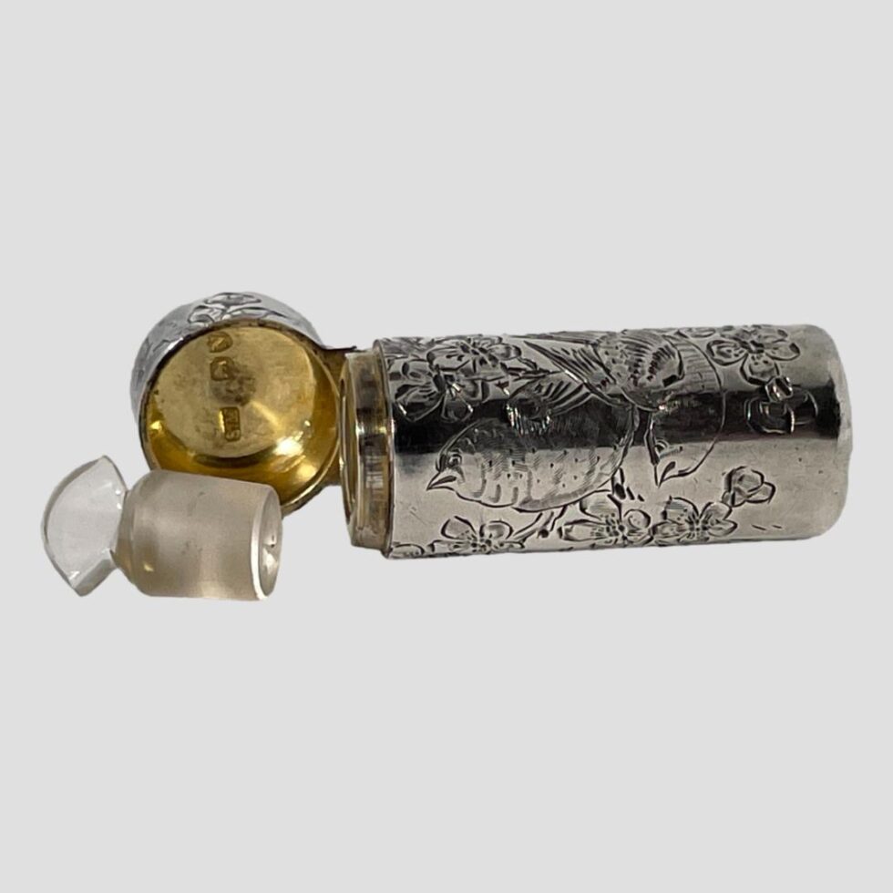 Photo of silver scent bottle shown open and on its side with the glass stopper removed and placed in front of the gilt interior of lid. The glass stopper is in very good condition with no damage. There is a short hallmark on the inside of the lid which is just visible.