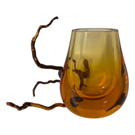 Main photo of Whitefriars amber hambone vase seen from an eye level and displayed with wider side forward facing. The vase called hambone as it resembles a ham bone. It has a thick rounded base with the orange colour darkest here before graduating to lighter shade at top. There is a dried twig behind the vase partially sticking out on the left.