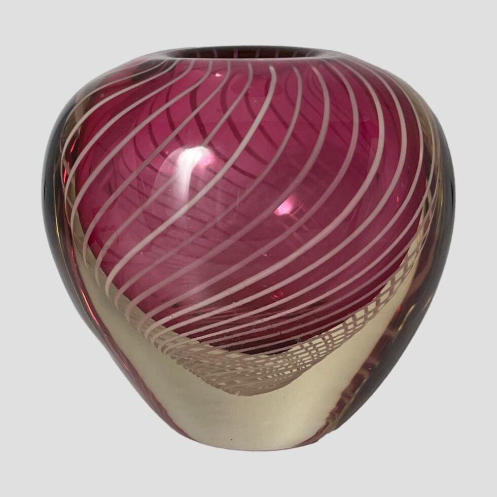 Main photo of Vicke Lindstand zebra vase seen from an eye level with one wide side on display. The shape of the vase is like a heart but without the little dip bit at the centre top. It has a narrower base in clear glass with some of the filigree strands looking like a fine net visible just below where the pink starts. The upper part balloons out into a bulbous finish with the vase opening sat in the middle at the top (not the whole of the top is open) The pink is a lovely shade and the white filigree swirls finish this piece perfectly.