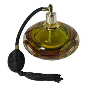 Main photo of modern Murano perfume atomiser displayed seen from a slightly raised angle looking diagonally downward. The glass bottle has 4 flat sides and is mainly a yellow/green with clear base and an amber sommerso inclusion. The black spray atomiser is to the left with the puffer ball and tassel in the left foreground. We are looking at the bottle with a none flat side in the foreground.