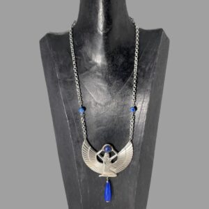 Main photo of Egyptian revival Isis necklace displayed on a black wooden stand with the necklace front forward facing. This stand is used for longer pieces and this necklace has a generous length with the goddess Isis taking centre stage of course! She is definitely a statement here She is kneeling with head and body facing right. Her arms are extended above her head where she holds a ball of lapis lazuli. The wings extend from behind the torso to the left and right and the tips of these are where the chain of the necklace is attached. 2 smaller lapis beads are added approximately 2 inches above the wing tips and then the chain continues all the way to the clasp. Below the goddess is a long water droplet shaped bead of lapis lazuli which hangs from the centre completing the look.